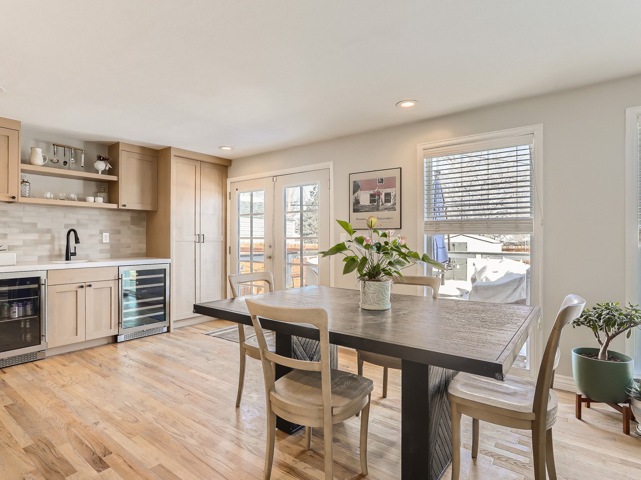 Bright dining area with a wooden table, four chairs, a plant centerpiece, and a kitchen adjacent. Large windows and glass doors let in natural light.