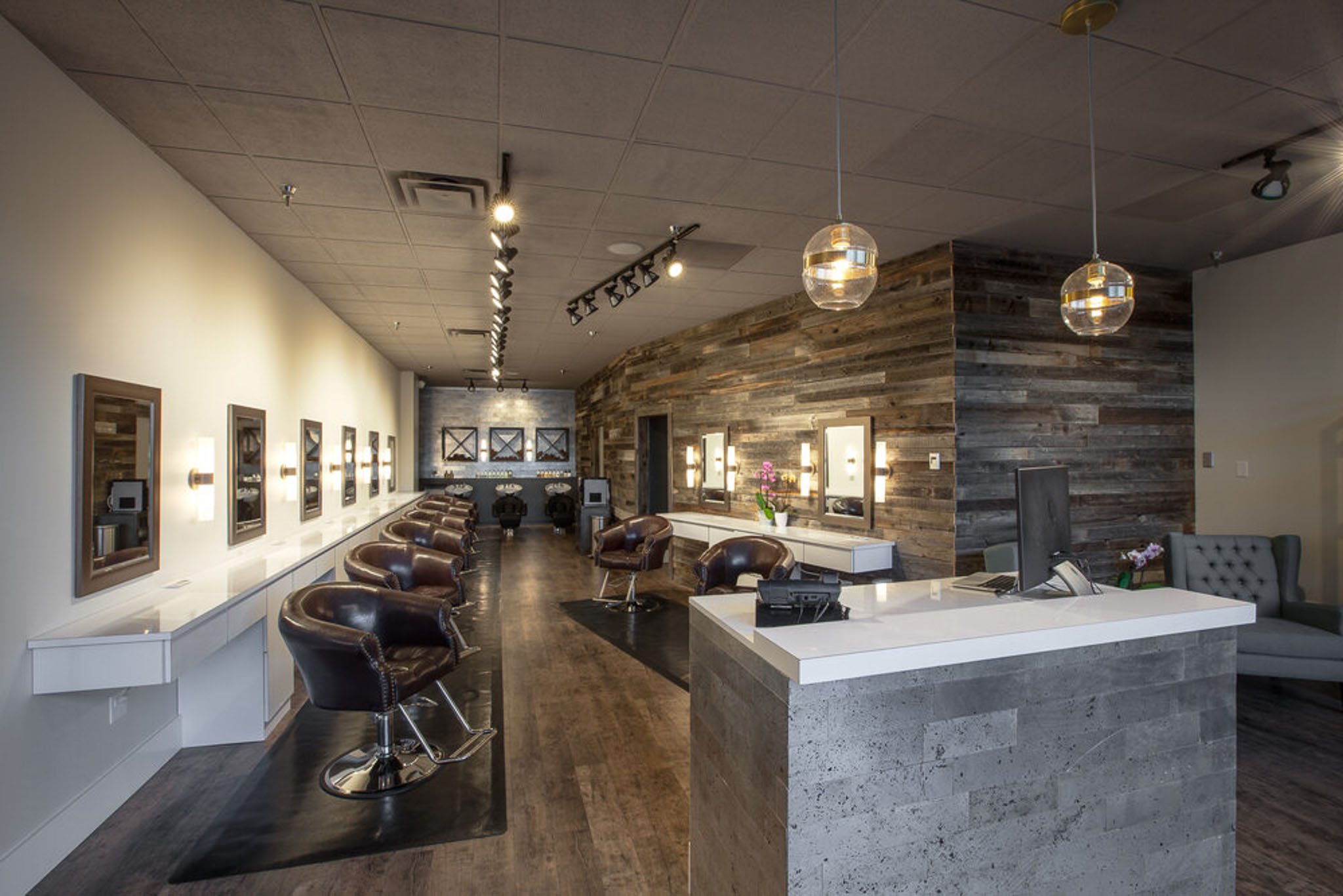 A modern hair salon interior with brown leather styling chairs, mirrors, and decorative pendant lights in a spacious, well-lit area. A reception counter is visible in the foreground.