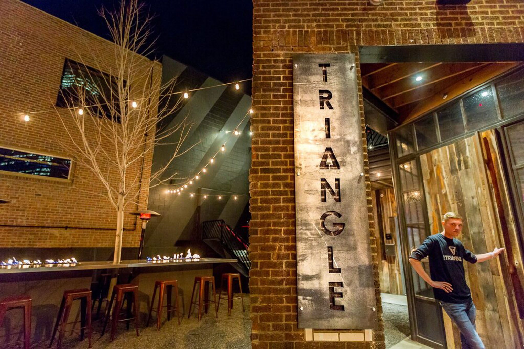 Outdoor bar with string lights, a long table with stools, and a large sign reading "TRIANGLE." A person is standing by the entrance.