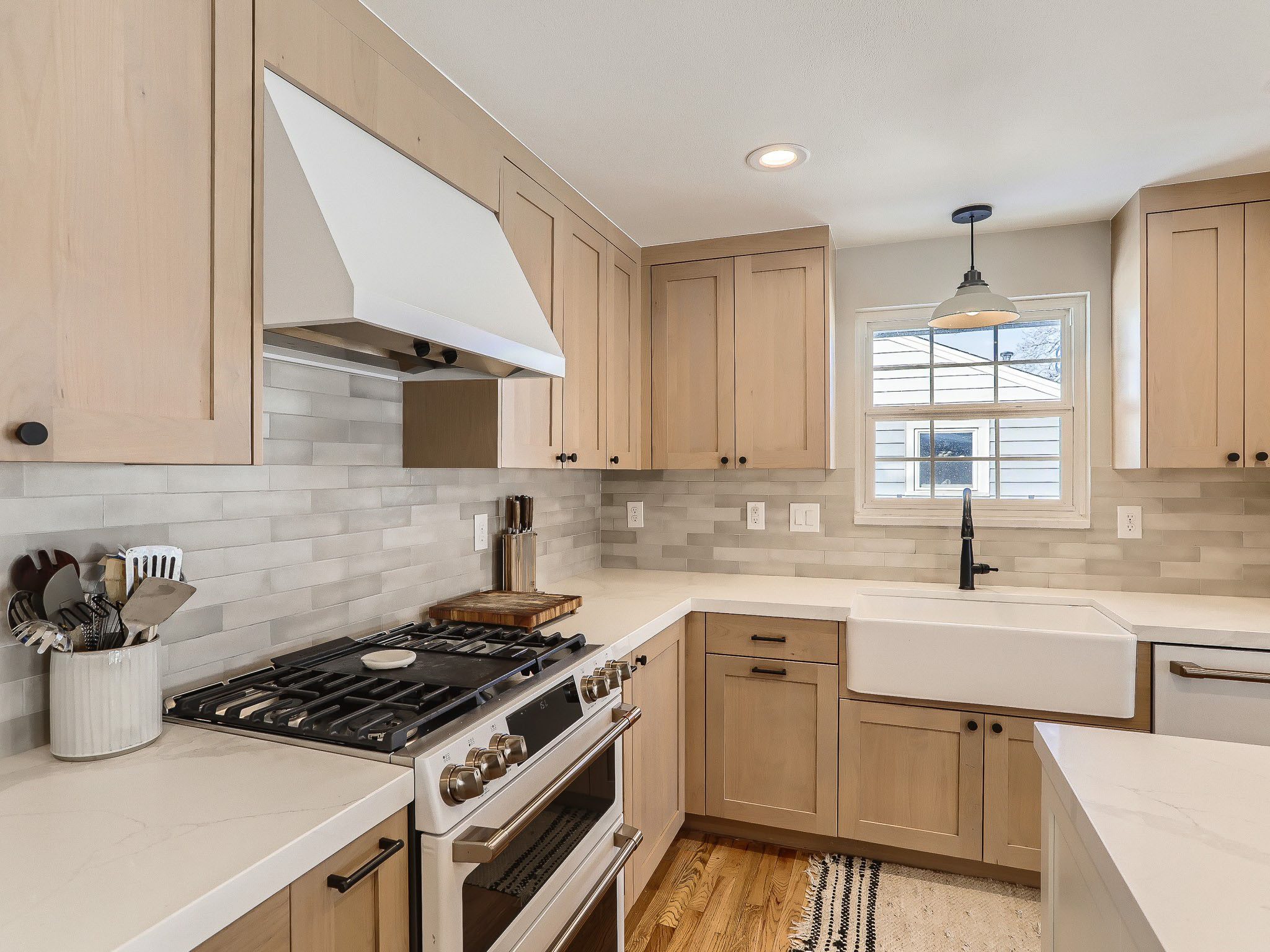 Modern kitchen with wooden cabinets, white countertops, stainless steel appliances, and a farmhouse sink. A window above the sink provides natural light.