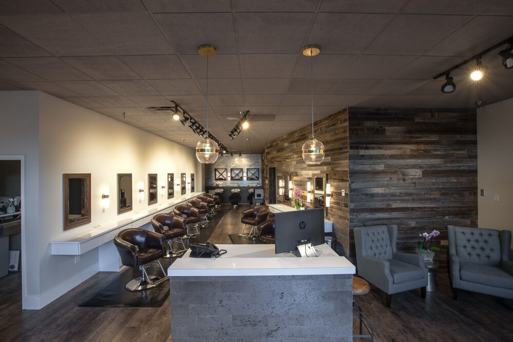 A modern hair salon with multiple styling chairs, a front desk, and a cozy waiting area featuring armchairs. The interior design includes wood-paneled walls and ambient lighting.