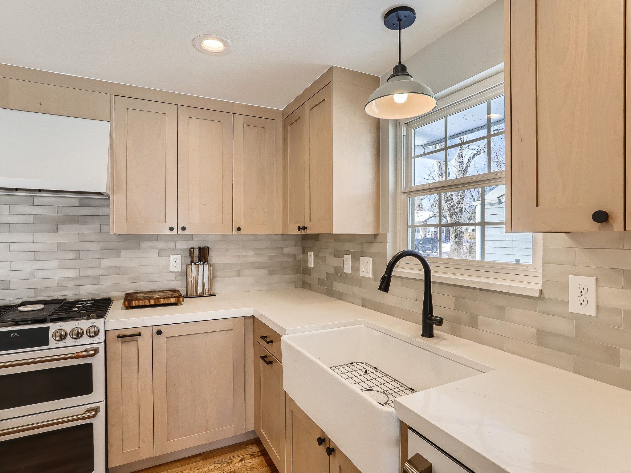 Modern kitchen with light wood cabinets, a white farmhouse sink, black faucet, subway tile backsplash, gas stove, and a window with natural light.