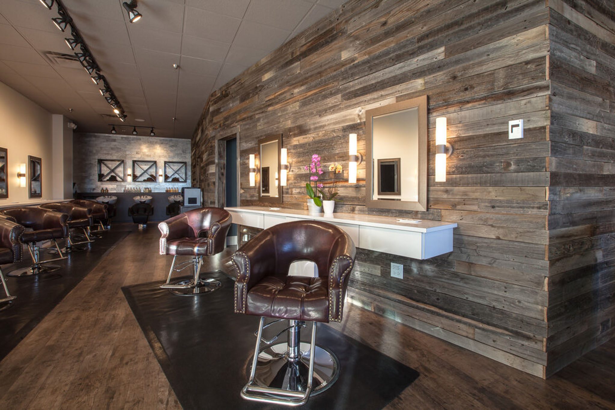 A modern salon with rustic wooden walls, leather styling chairs, and mirrors. The room has overhead track lighting and a vase with orchids on a counter.