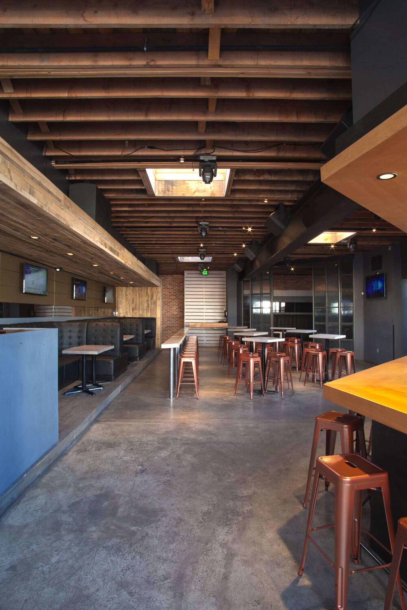 Modern, industrial-style bar interior with high ceilings, exposed beams, metal stools, and wooden tabletops. Large windows allow natural light; multiple TVs are mounted on the walls.