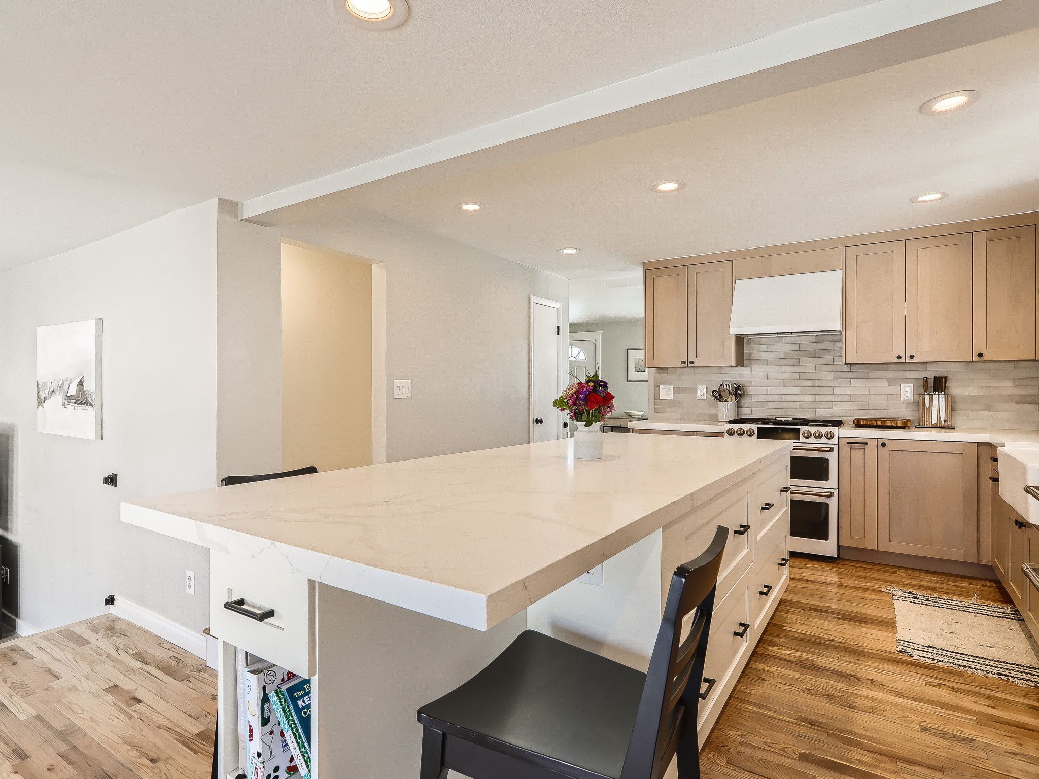 Modern kitchen with wooden cabinets, marble island, and light hardwood floors. Black chairs at the island and a vase with flowers are on it. Walls are white with recessed lighting.