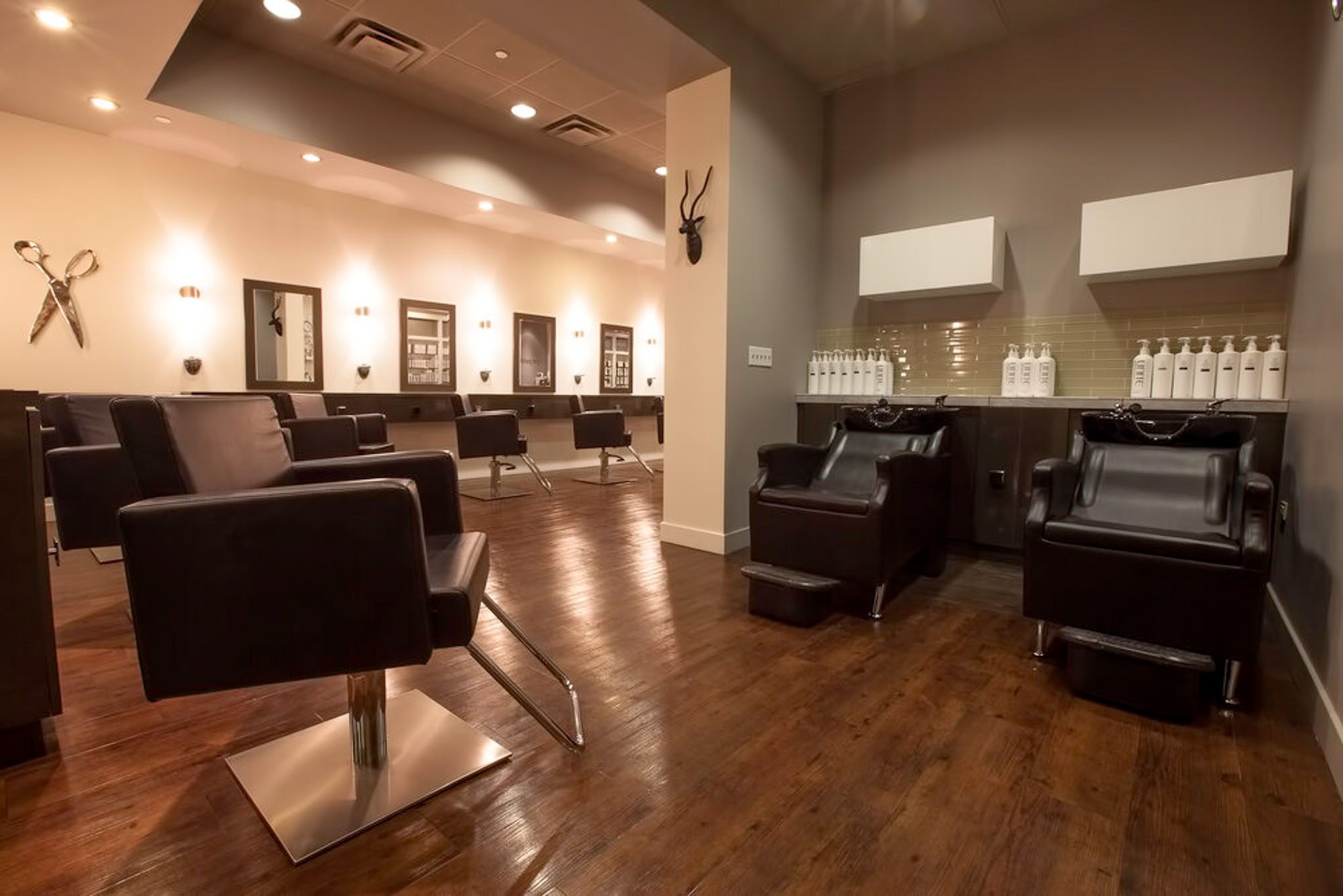 A modern hair salon with empty black chairs, styling stations with mirrors, and shampoo basins on a wooden floor.