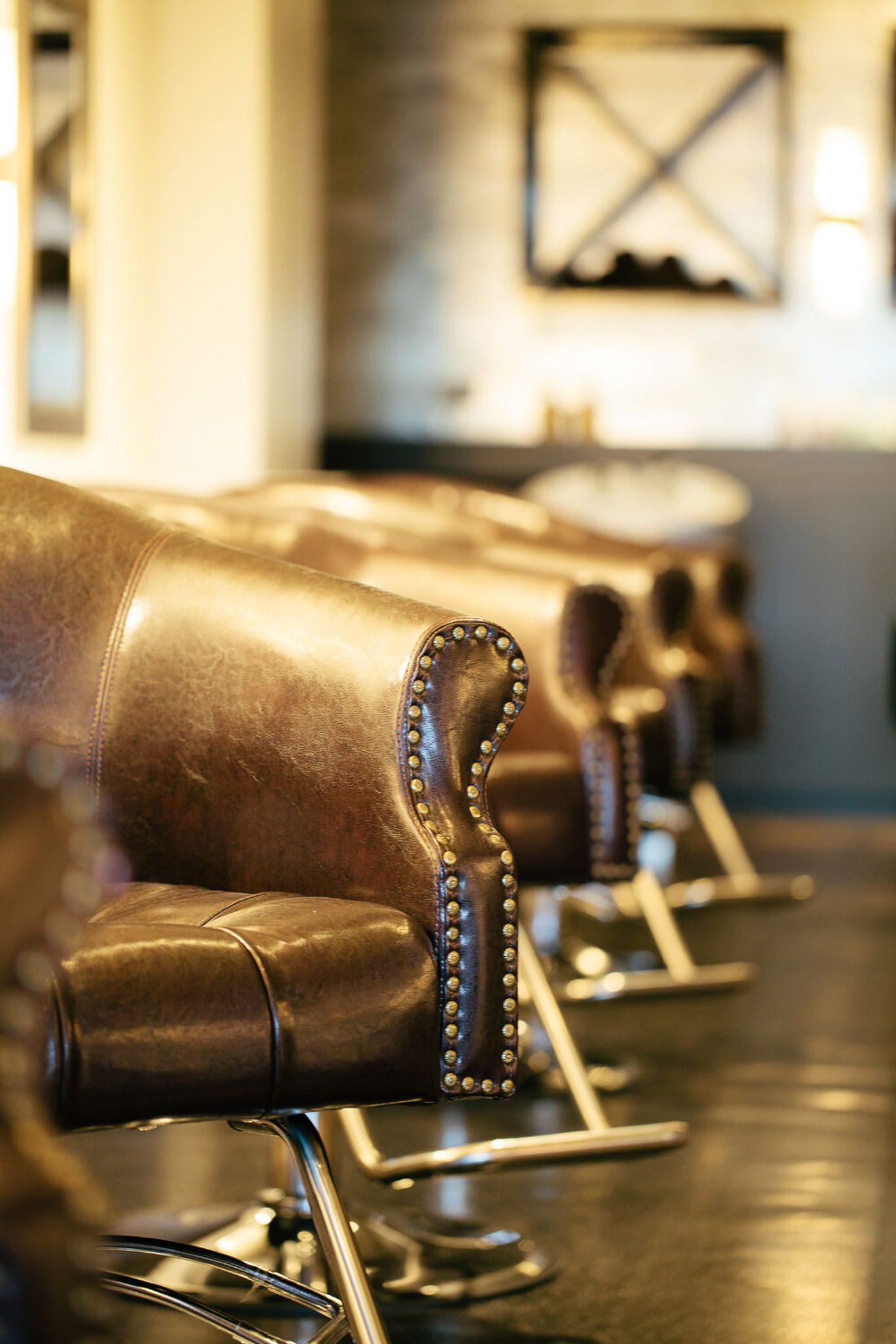 Row of brown leather salon chairs with metal studs, lined up in a dimly lit space with blurred background.