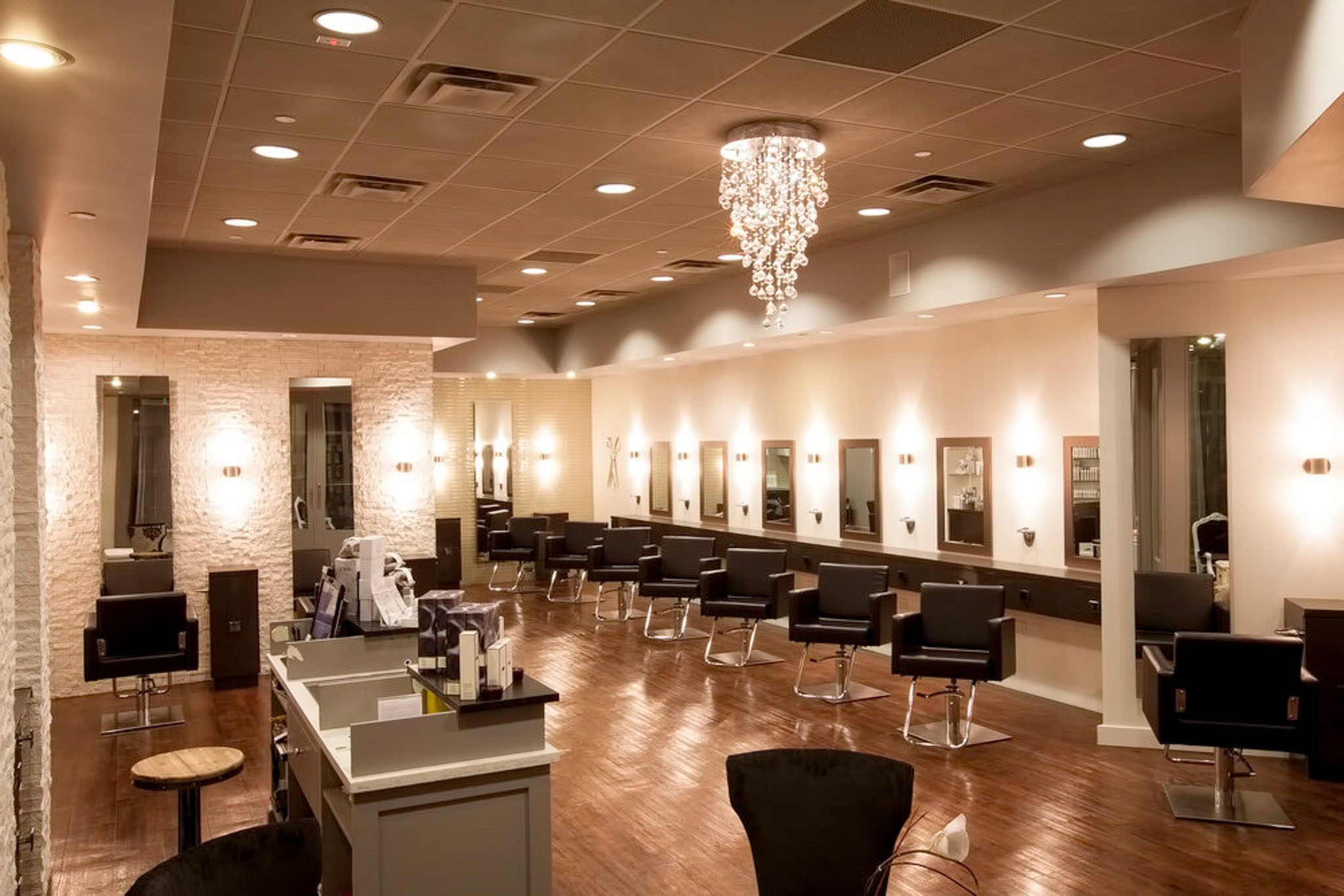 Modern hair salon interior with black styling chairs, large mirrors, and a central crystal chandelier on a wooden floor.