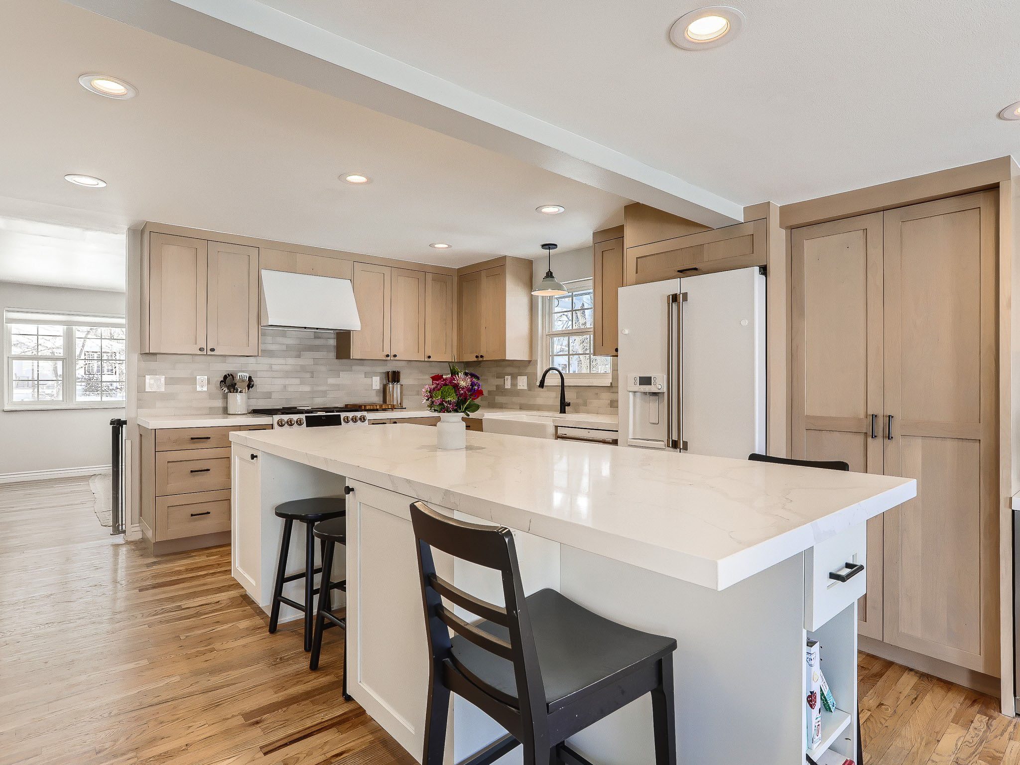 Modern kitchen with light wood cabinets, white countertops, an island with chairs, and a vase of flowers. The room has hardwood floors and stainless steel appliances.
