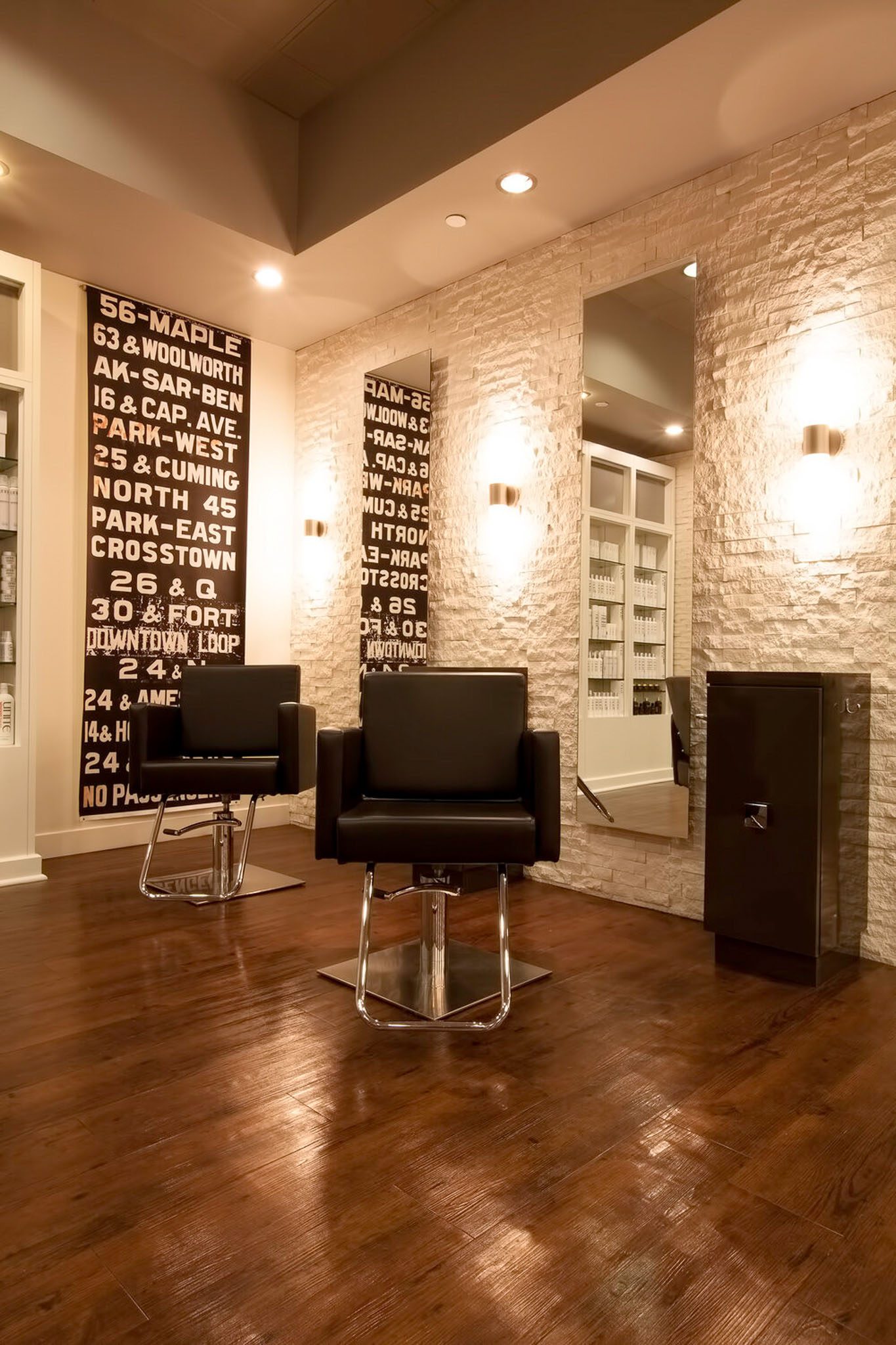 A modern hair salon with two black salon chairs, mirrors on a brick wall, and a wooden floor. Urban-themed decor is displayed on wall panels.