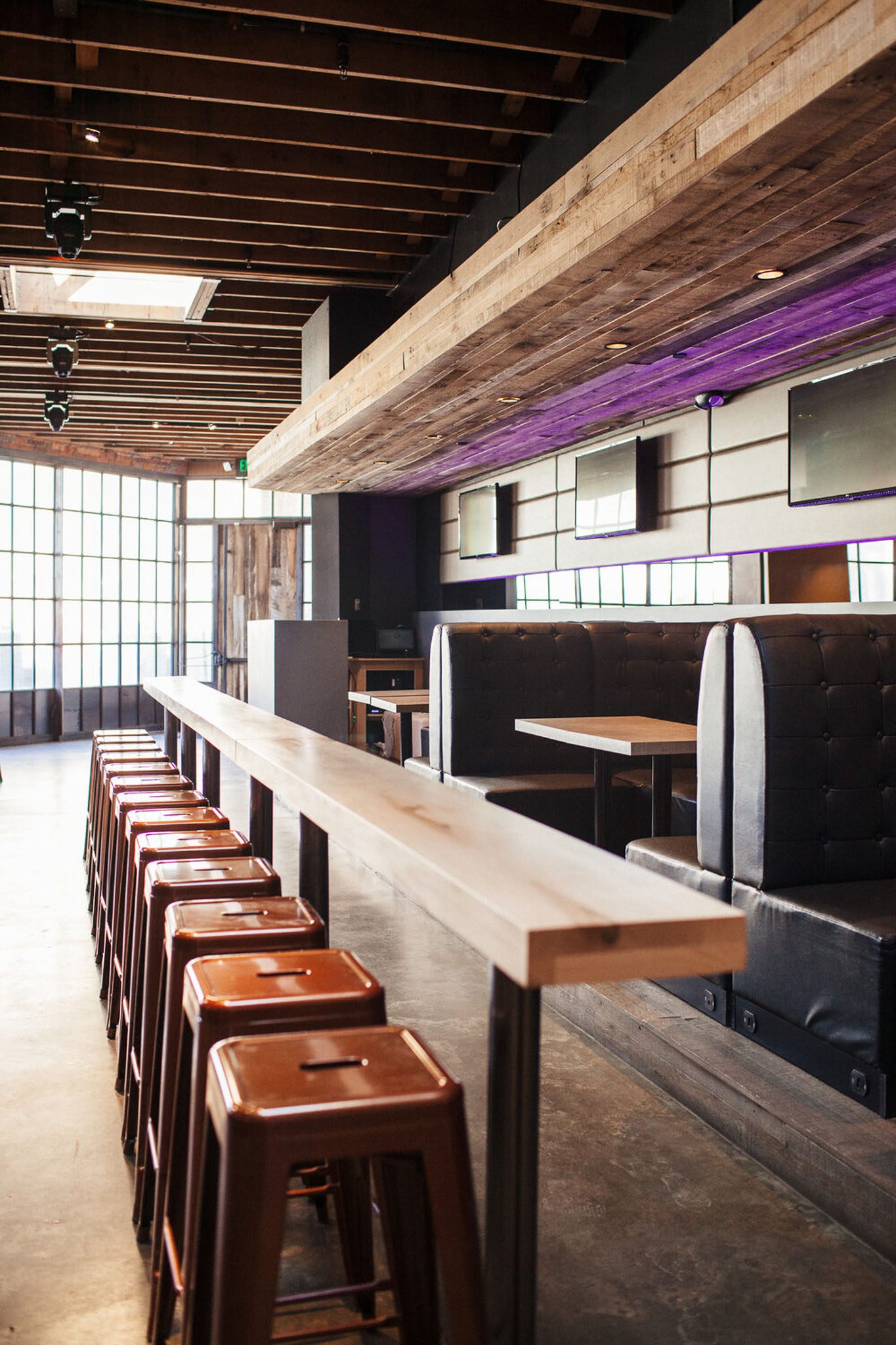 Empty modern bar interior with high wooden bar, metal stools, booth seating, and TV screens on walls, featuring exposed wood ceiling and large windows.