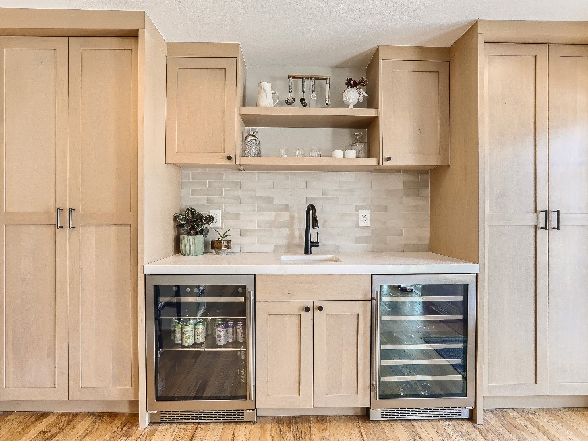 A kitchen area with light wood cabinets, a white countertop, a black faucet, a tile backsplash, and two under-counter glass-front refrigerators.