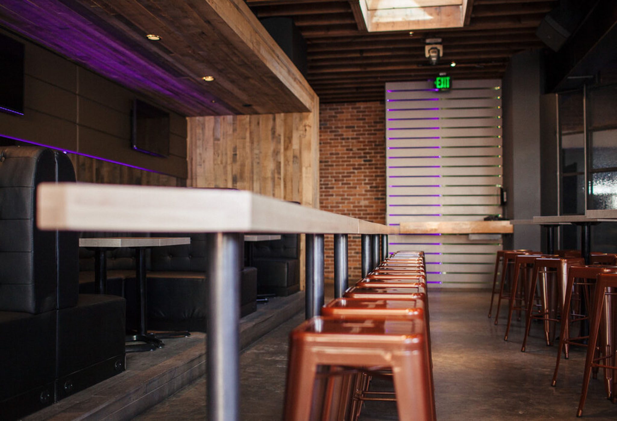 Industrial-style bar interior with wooden accents, high metal stools, and exposed brick walls. Long counter stretches through the space with muted lighting from ceiling fixtures.