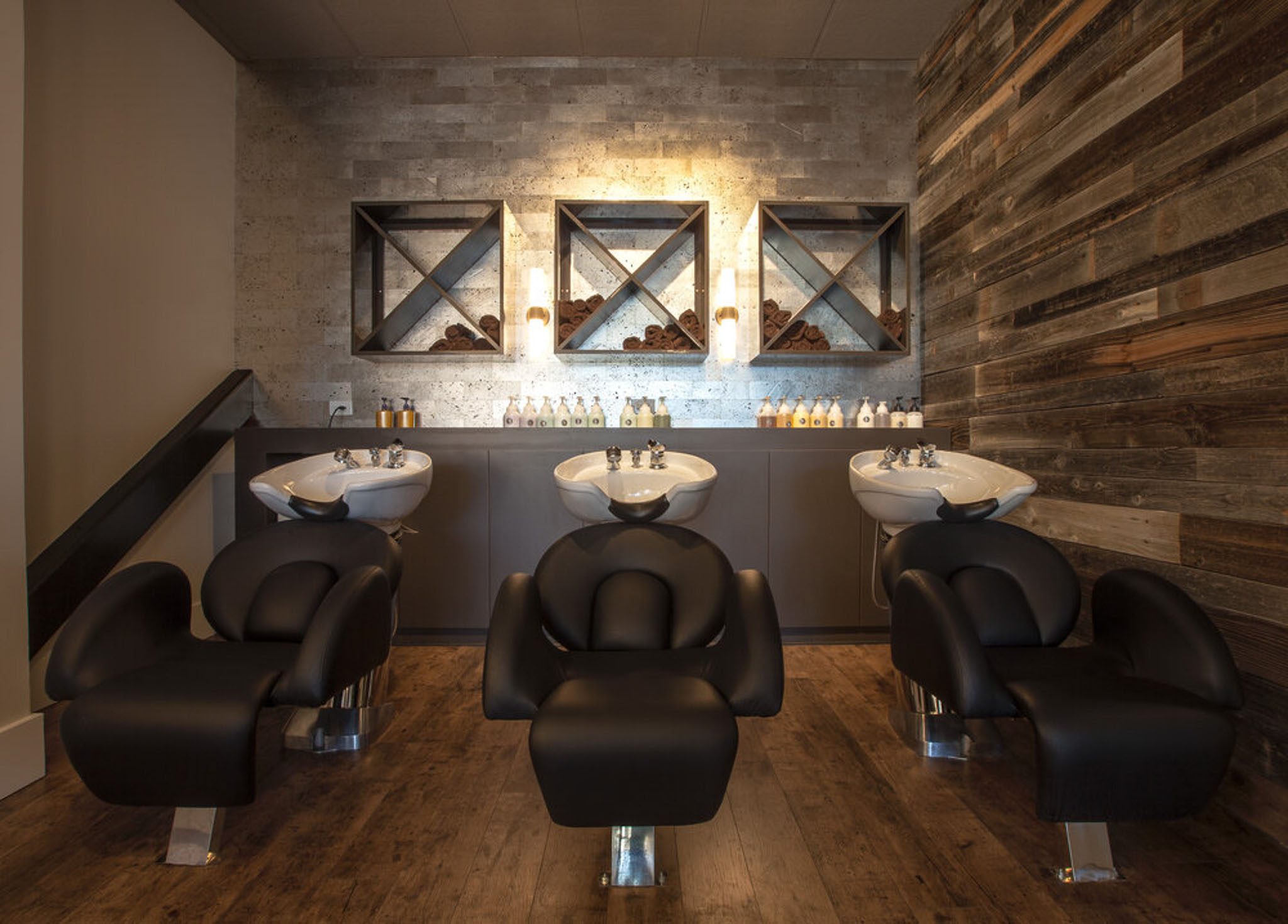 Modern salon interior with three black hair-washing stations, wooden flooring, and decorative shelving on walls.