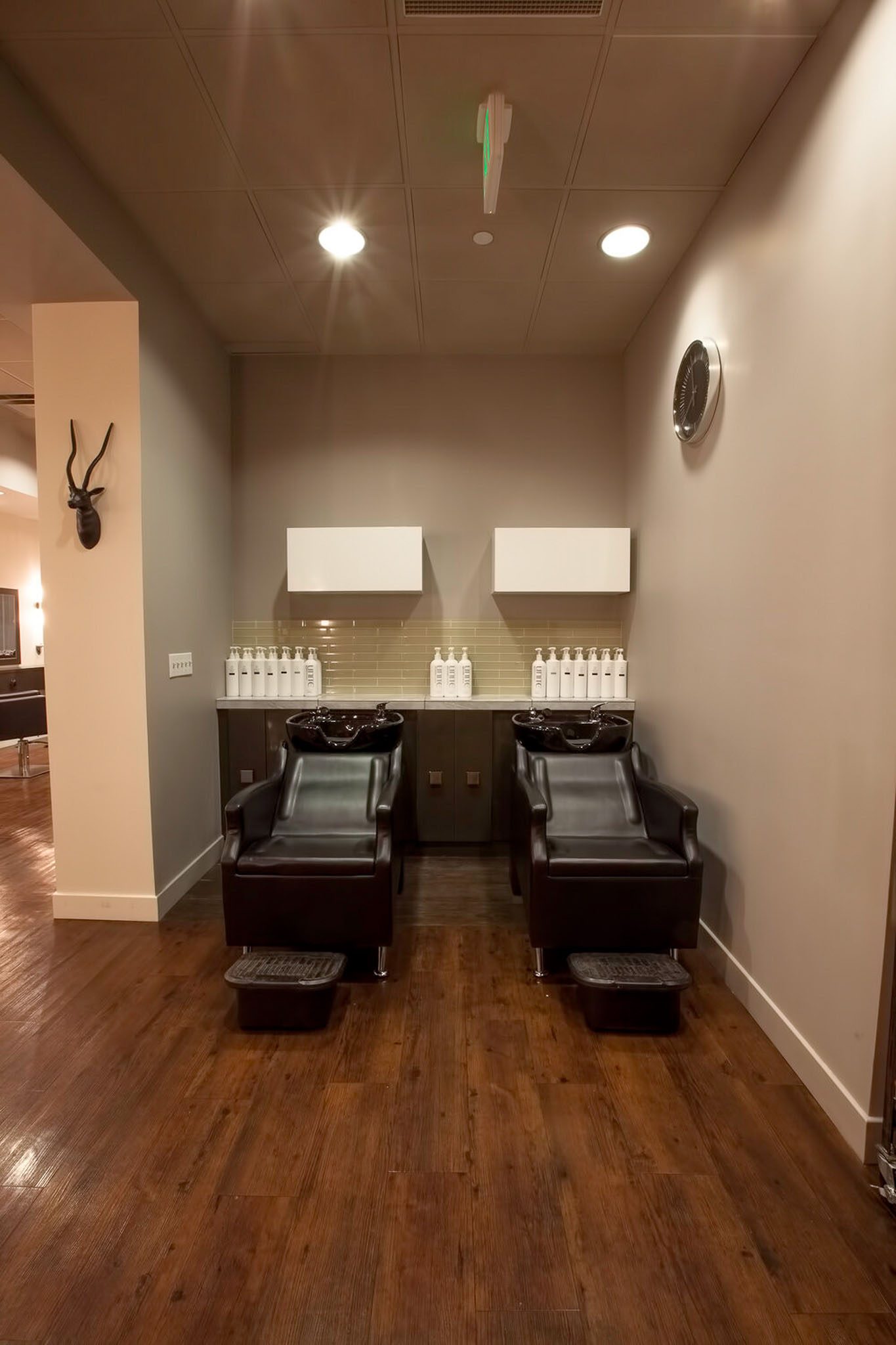 Two black salon chairs with shampoo sinks in a well-lit room. White bottles line the shelves above, and a clock is on the wall.