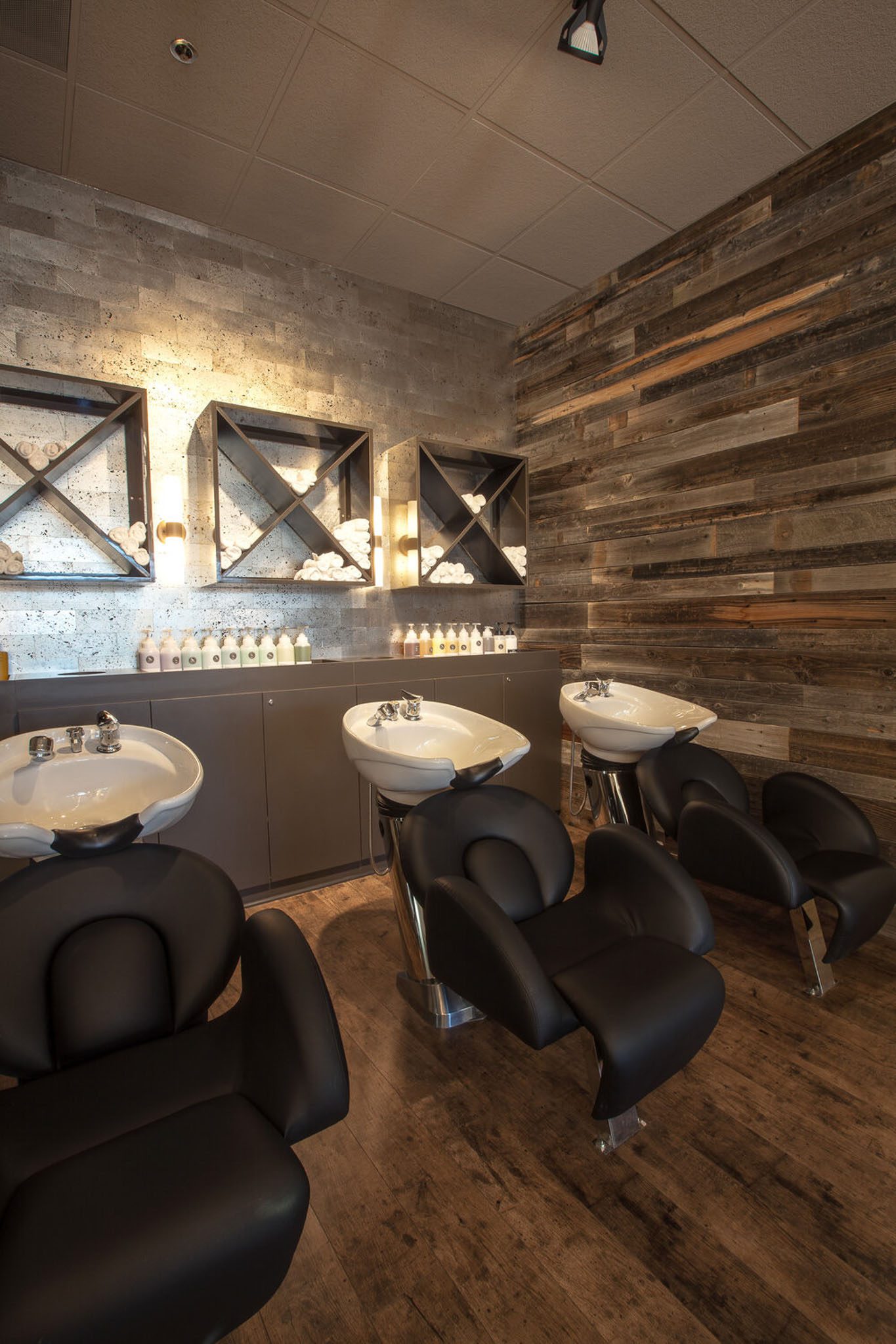 Hair salon interior with three black shampoo chairs and sinks. Shelves with hair products are mounted on a textured wall with warm lighting. Wooden floor and walls add a rustic touch.