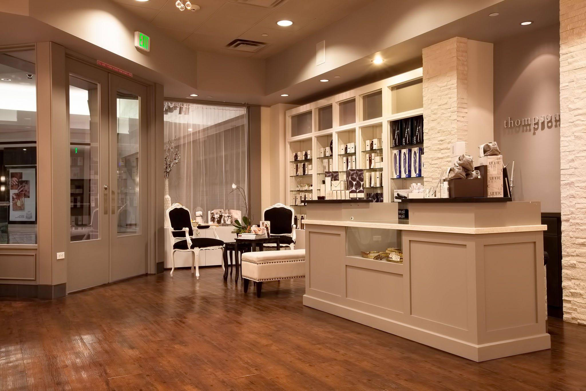 A modern boutique interior with a wooden floor, gray walls, and a reception area. Shelving with products is in the background, alongside a small seating area with white chairs and a table.