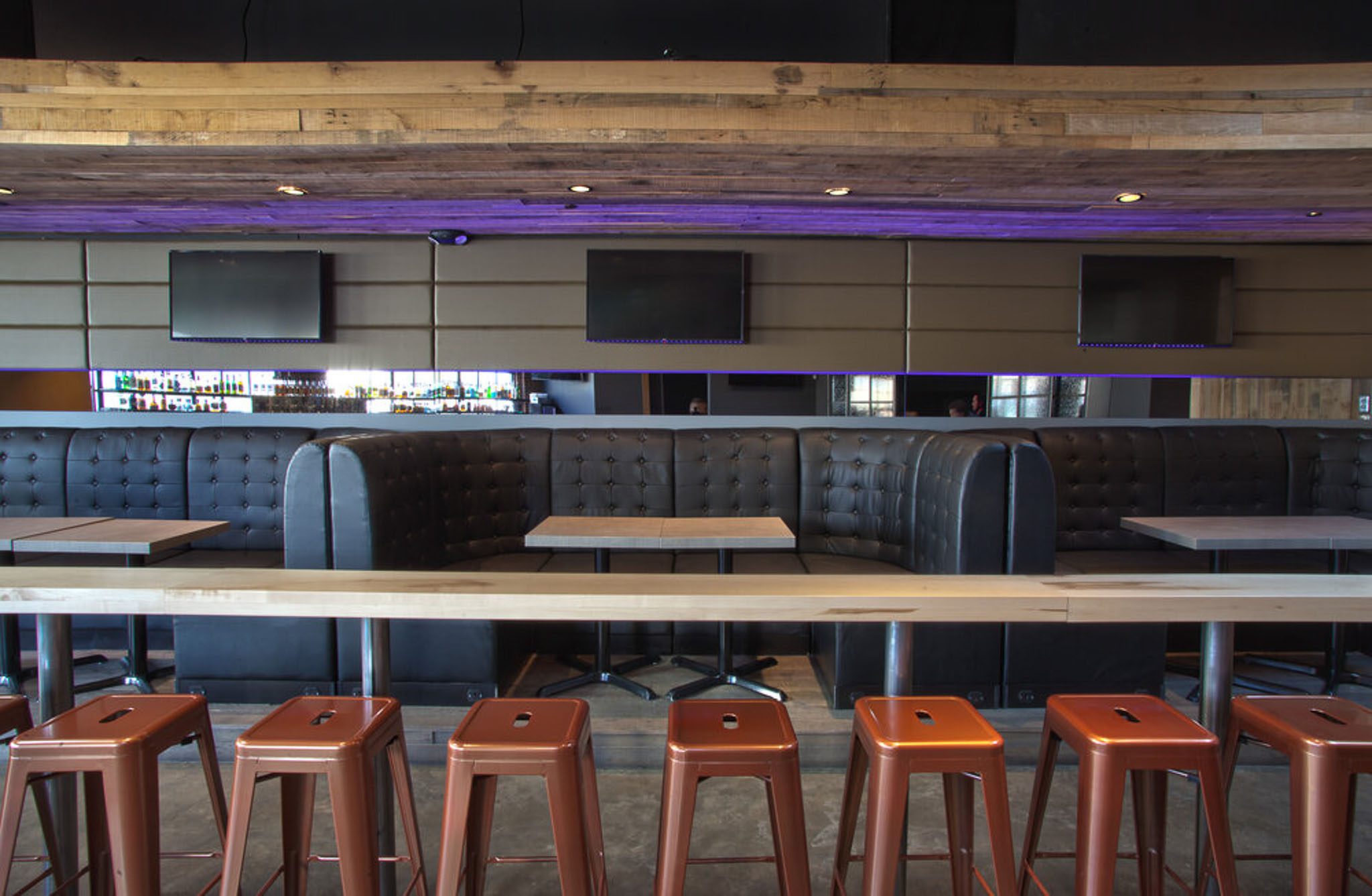 Empty modern bar with brown leather booths, wooden tables, and a row of red metal stools. Three blank TVs are mounted above.
