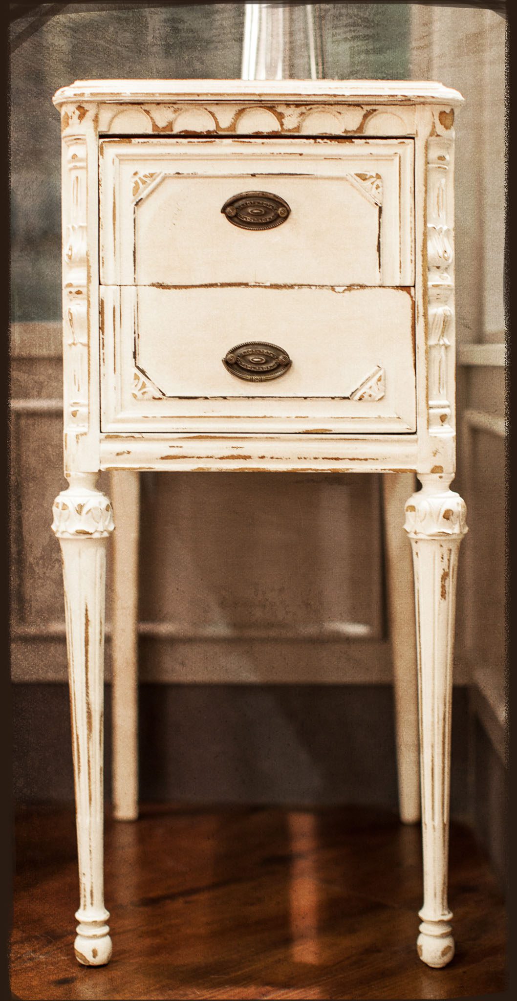 A worn, vintage-style wooden nightstand with two drawers and ornate legs, painted white with a distressed finish.