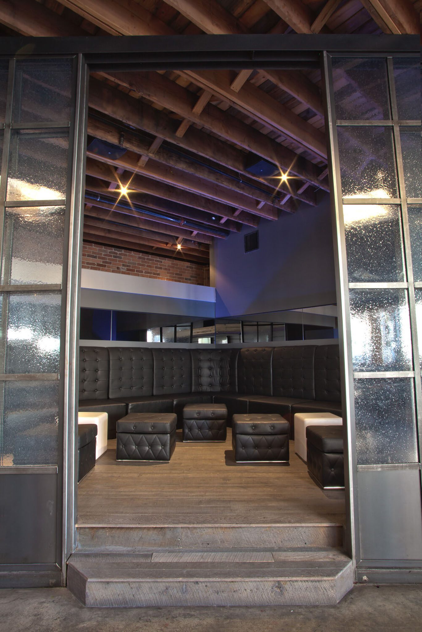 A modern lounge with black cushioned seating, square ottomans, and a wooden ceiling, framed by glass doors.