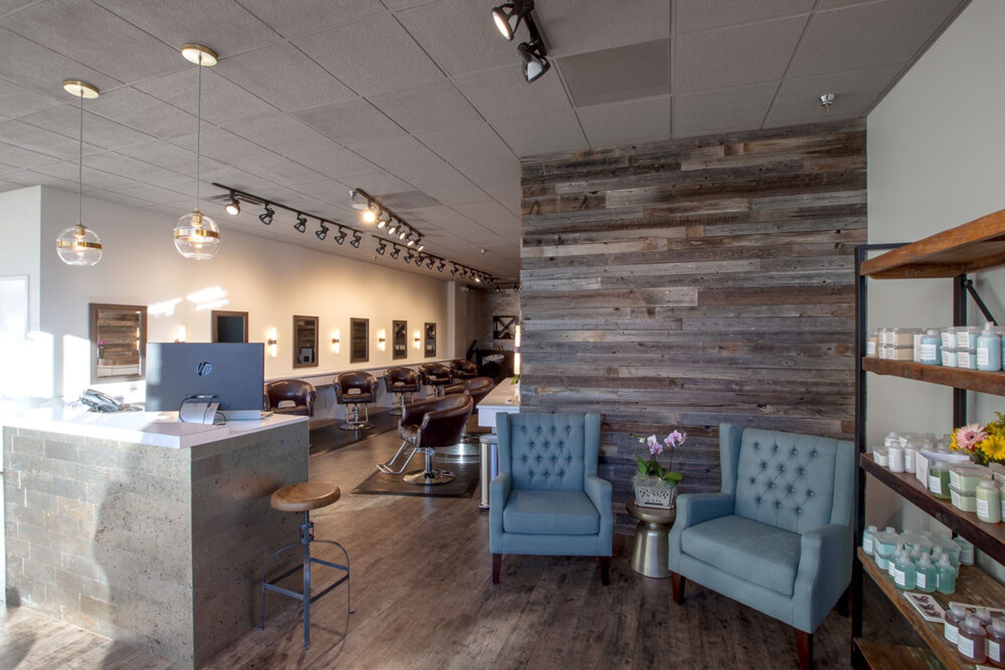 Hair salon interior with rustic wood accents, modern lighting, blue armchairs, and a front desk. Shelves display hair products, and there are styling stations in the background.