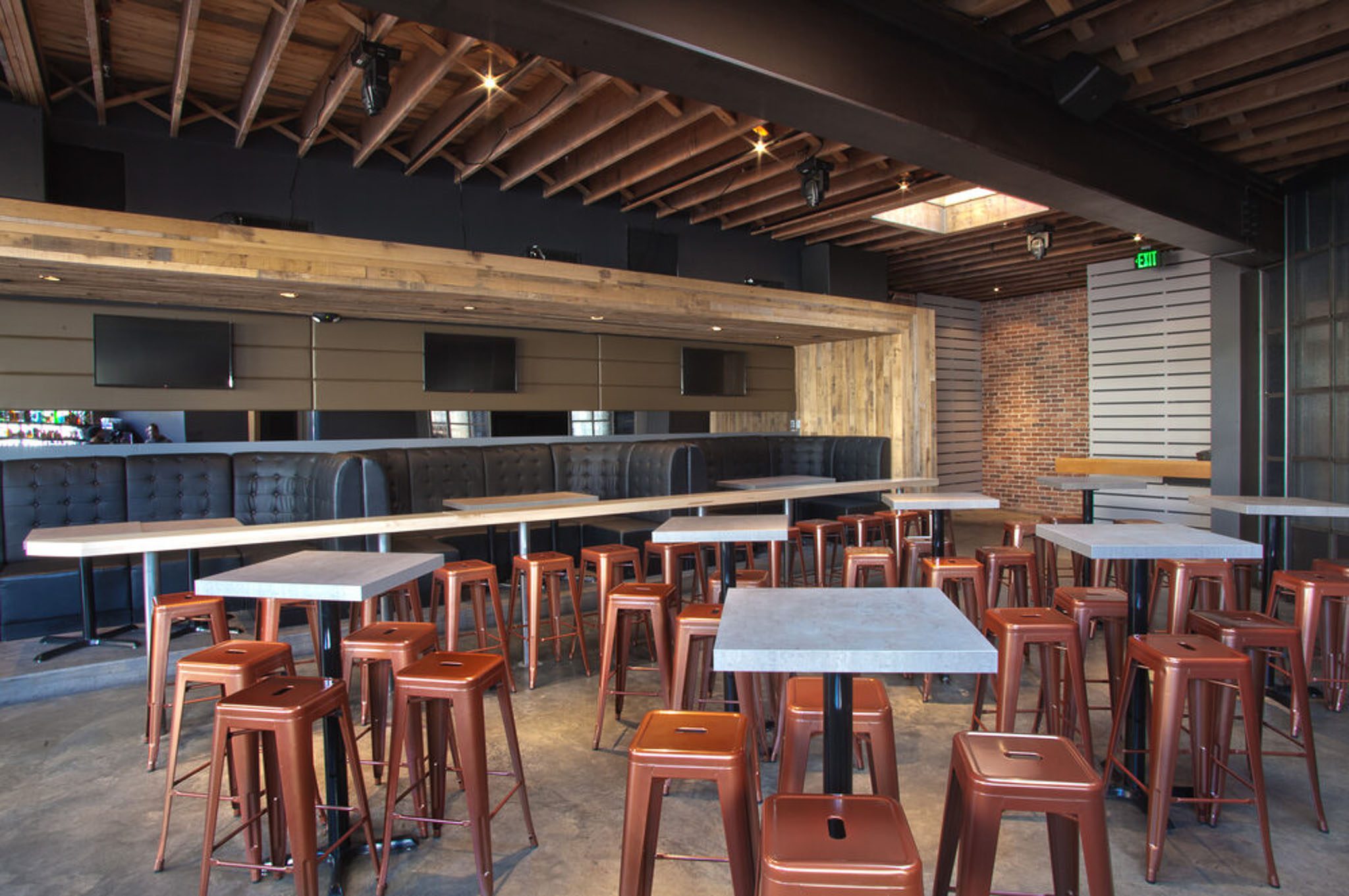 A modern bar interior with concrete tables, copper stools, and exposed wooden beams. The space features a mirrored wall, a brick wall, and a wooden ceiling.
