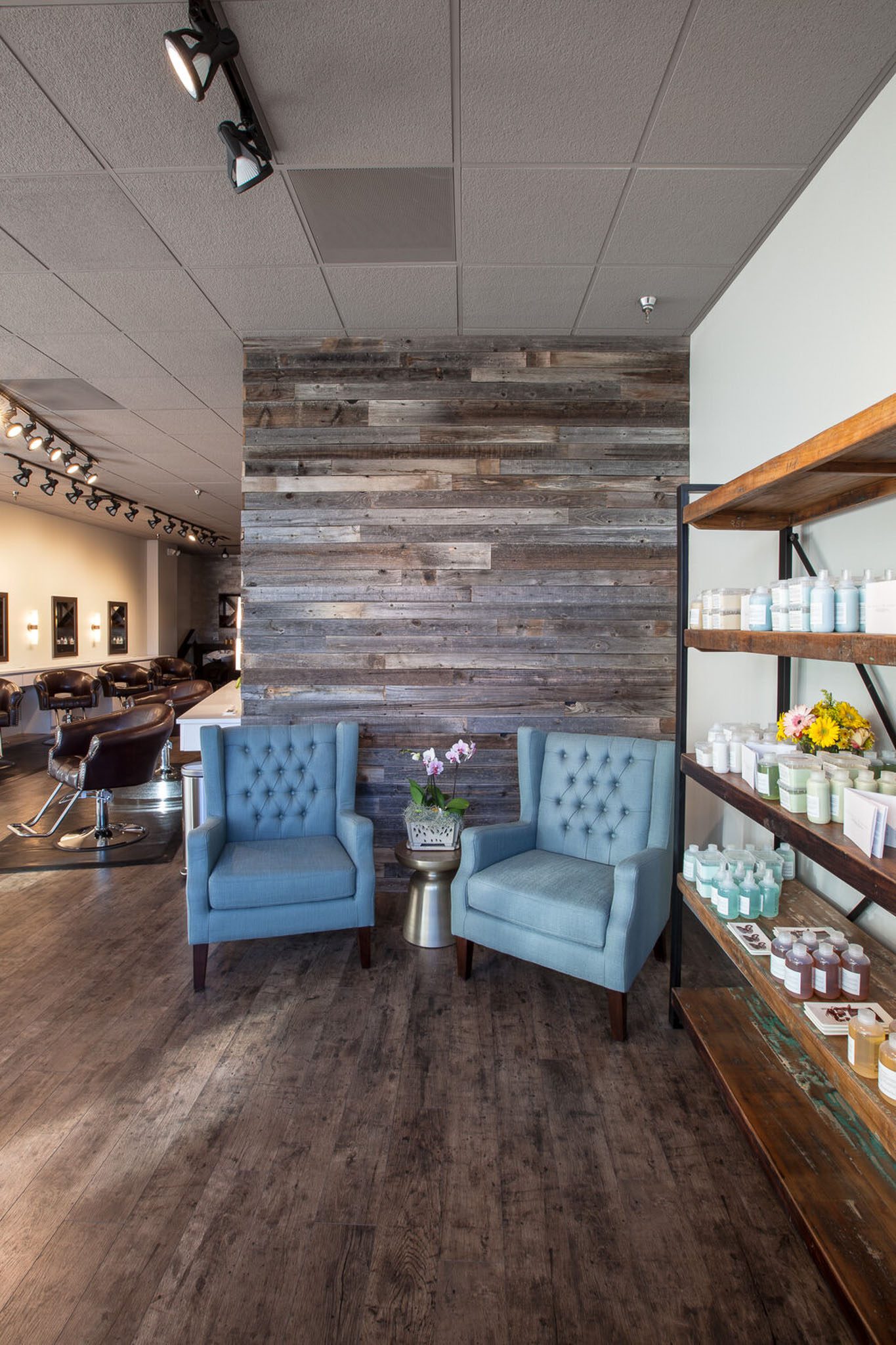 A salon interior with two blue chairs and a small table with flowers. A wooden wall is behind the chairs, and shelves with hair products are on the right. Haircutting stations are visible in the background.