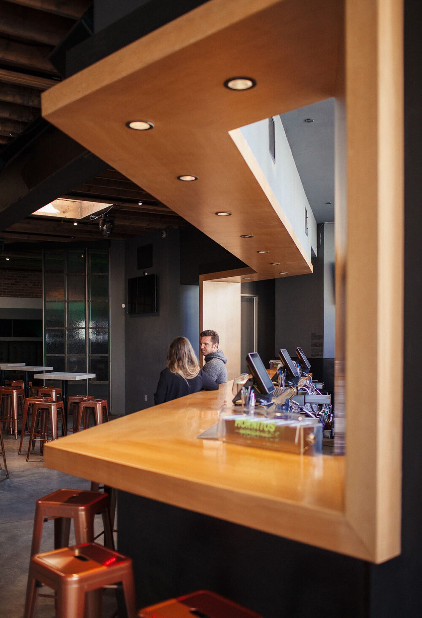 Interior of a modern bar with wooden counter and stools. Two people sit in conversation at the bar. Ceiling lights and a menu holder are visible.