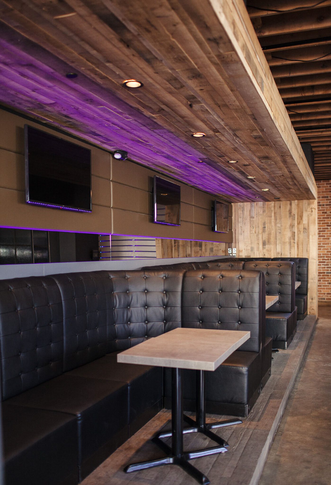 A modern bar interior with black leather booth seating, a wooden table, multiple wall-mounted screens, and a wooden ceiling with purple lighting.