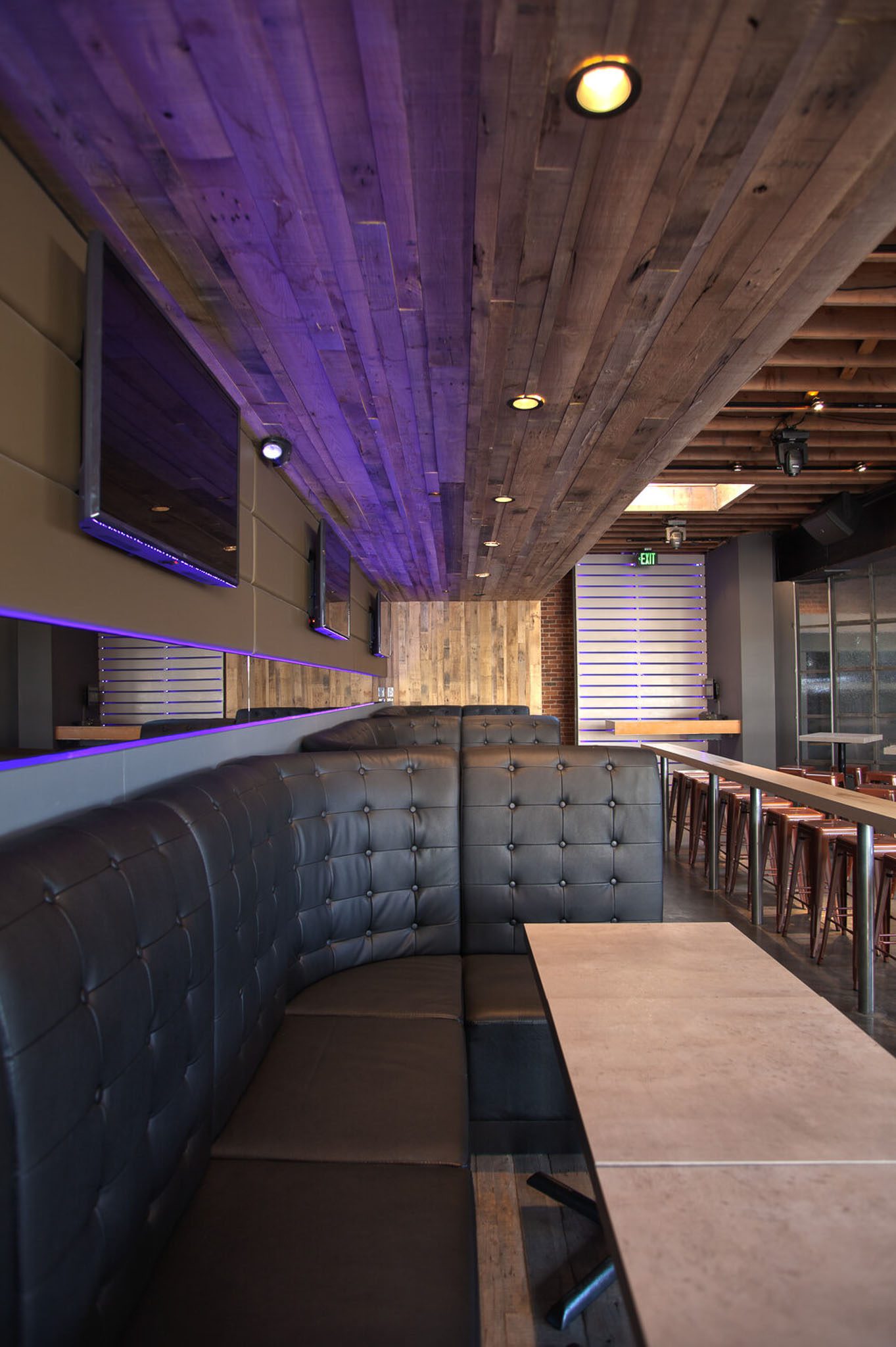 Interior of a modern restaurant with black tufted booth seating, wooden ceiling, mounted TVs, and high tables with stools. The setting is dimly lit with ambient purple lighting.