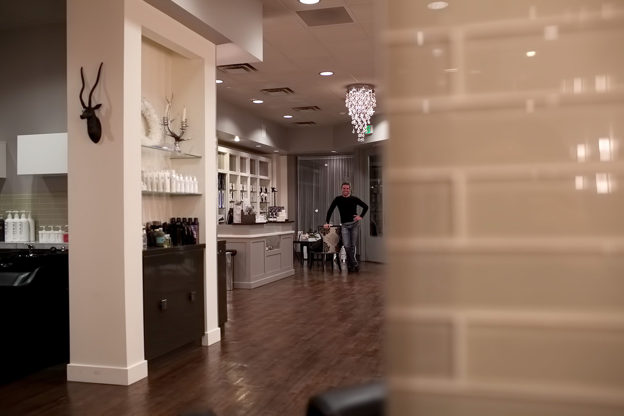 A man stands in a well-lit salon with wooden floors, surrounded by shelves of hair products and a stylist chair. A decorative light fixture and wall-mounted antelope head are visible.