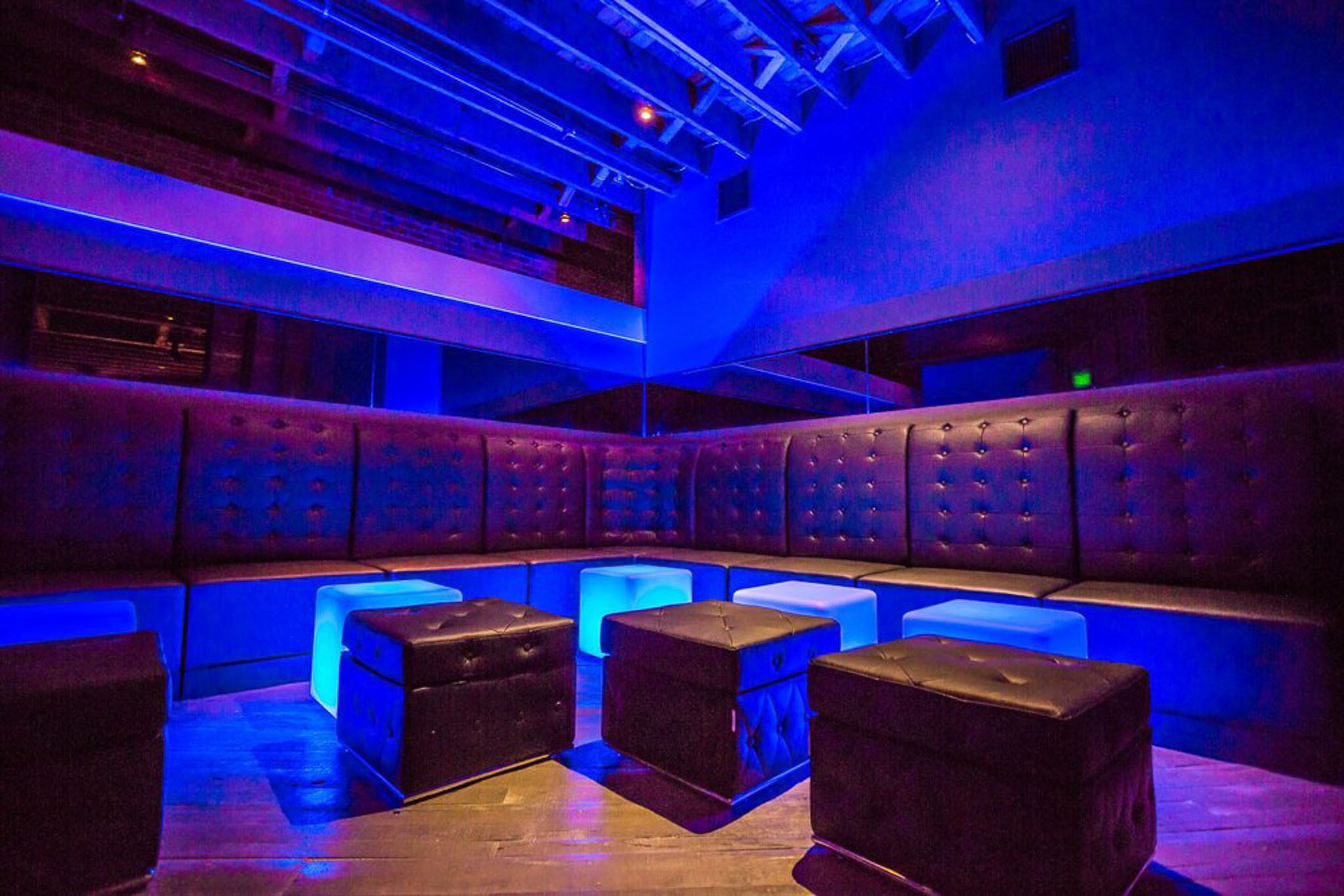 A dimly lit lounge with blue lighting, featuring tufted black seating along the walls and cube-shaped ottomans.