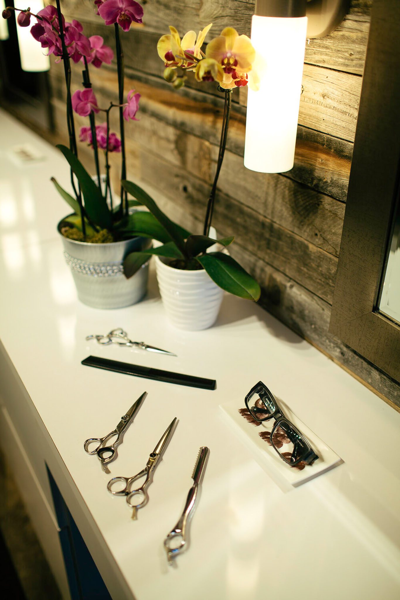 A white countertop with hairdressing scissors, a comb, eyeglasses, and potted orchids in the background.