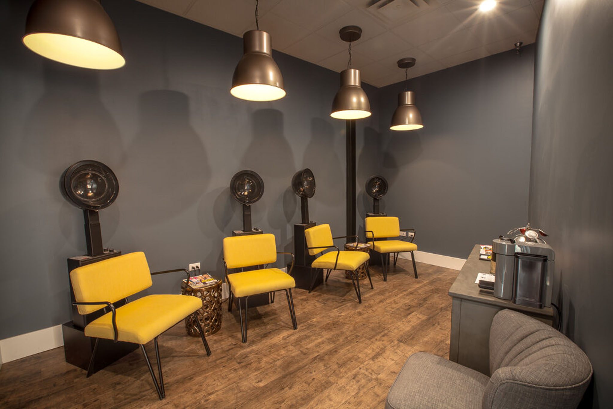 A modern salon waiting area with four yellow chairs, hair dryers, dark walls, and overhead pendant lights. A gray armchair and coffee machine sit on the right.