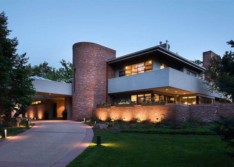Modern two-story house with brick facade, large windows, and illuminated pathway, set against a twilight sky.