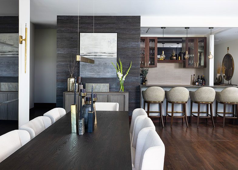 Modern dining room with a dark wood table, light gray chairs, and decorative bottles. A bar area with stools is in the background. Walls are dark textured with artwork and sconces.