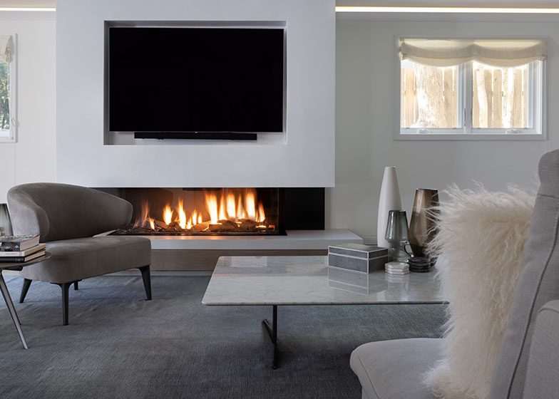 Modern living room with a wall-mounted TV above a linear fireplace, gray chairs, and a marble coffee table. Soft lighting from a window illuminates the space.