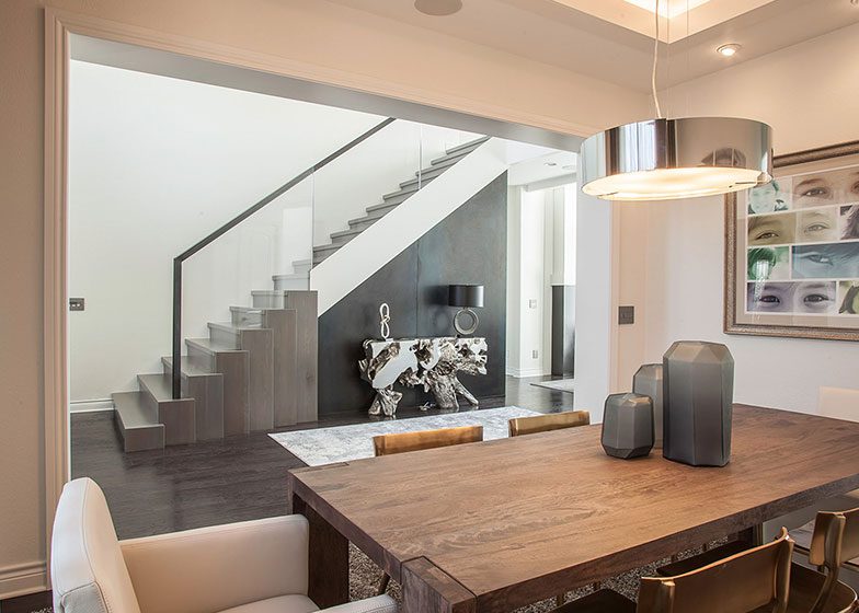 Modern dining area with a wooden table, white chairs, and a pendant light. A staircase with glass railing and a decorative console table is visible in the background.
