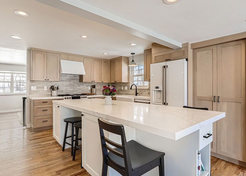 Spacious kitchen with light wood cabinets, a large white island countertop, black chairs, and stainless steel appliances. A vase with flowers is on the island. Natural light fills the room.