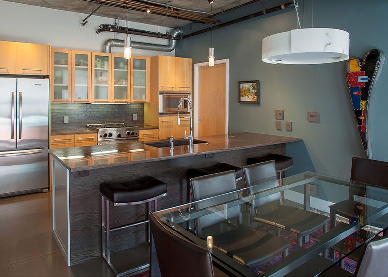 Modern kitchen with wooden cabinets, stainless steel appliances, and a central island with bar stools. A glass dining table is in the foreground, and a snowboard decorates the wall.