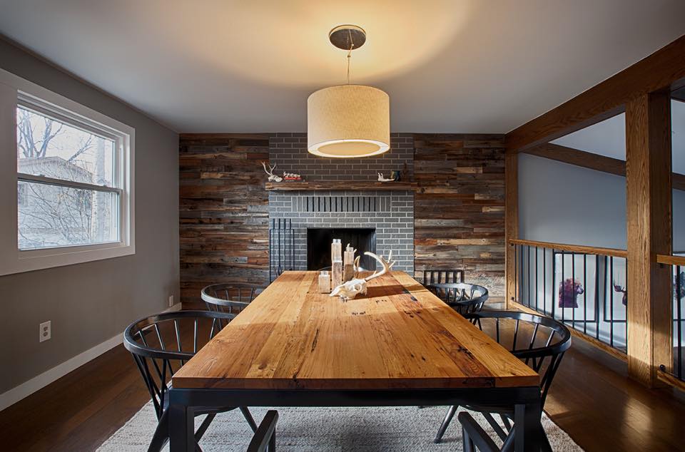 A cozy dining room featuring a wooden table, black chairs, a pendant light, and a fireplace set in a brick and wood accent wall. Natural light enters through a window on the left.