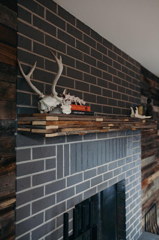 A dark brick fireplace with a wooden mantle features a pair of antlers, a skeletal bone, and stacked books.