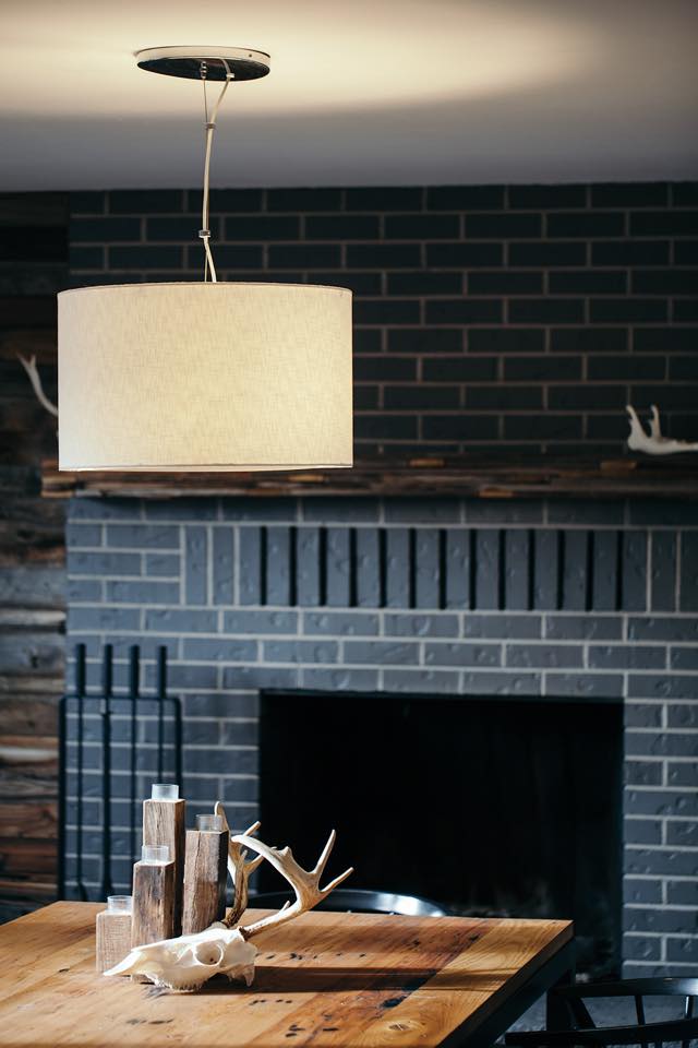 A modern dining area with a wooden table, antler decor, a large pendant light, and a brick fireplace in the background.