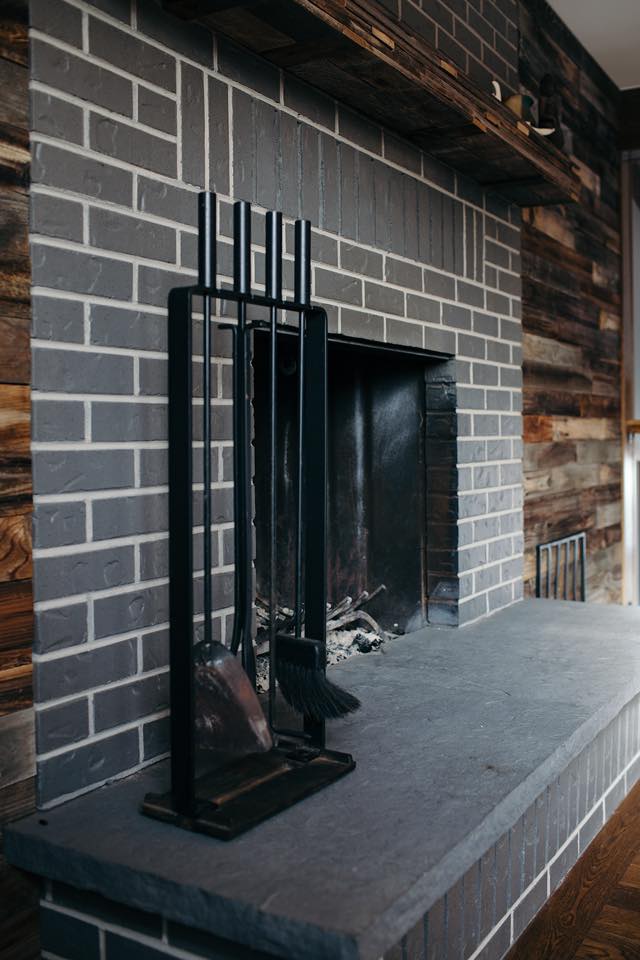 A brick fireplace with a dark interior, flanked by rustic wood paneling. Fireplace tools are placed on the stone hearth.