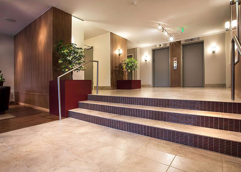 A well-lit lobby with tiled floors, stairs, and two elevators. Two large potted plants are placed on each side near the elevators.