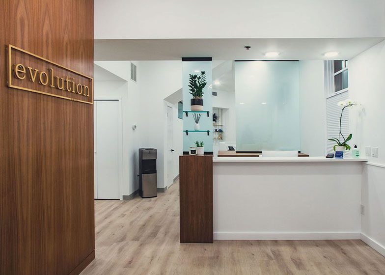 Modern reception area with wooden flooring, white walls, a desk, plants on glass shelves, and a sign saying "evolution" on a wooden panel.