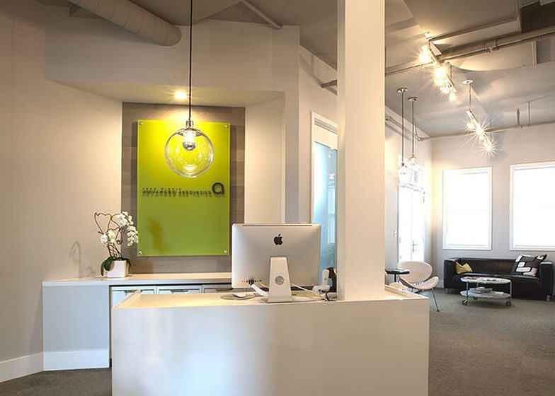 Modern dental office reception with a sleek white desk, an iMac computer, a green sign on the wall, and a seating area with contemporary lighting.