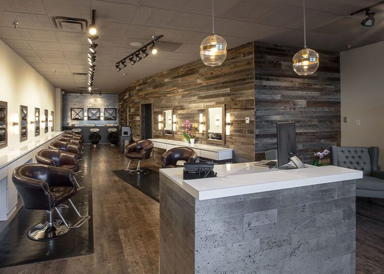 Modern salon interior with brown leather chairs, wooden accent walls, a white reception desk, and pendant lights. Mirrors line the left wall, and a waiting area with gray seating is on the right.