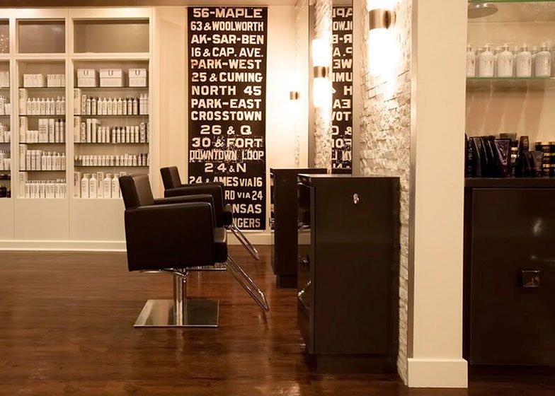 Interior of a modern salon with black chairs, mirrored stations, product shelves, and rustic wooden flooring. Bold wall art with text decorates the space.