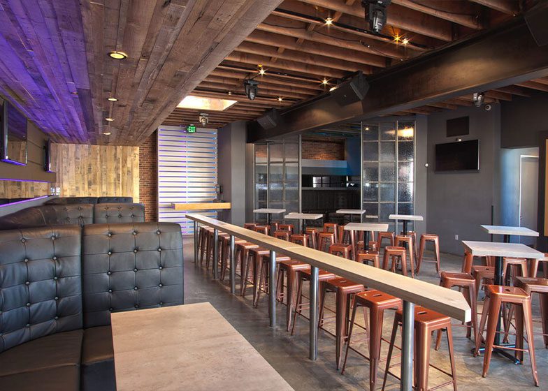 Modern, empty bar interior with wooden ceiling, tables, and stools. Black leather booths line one side, and the space is dimly lit with purple accents.